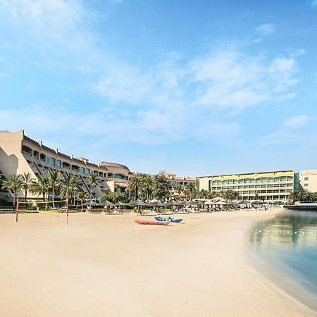 Al Raha Beach Resort & Spa Abu Dabi Exterior foto The photo depicts a serene beach scene with a clear blue sky. In the foreground, there is a sandy beach that gently slopes down to calm water. On the left side, several palm trees provide a tropical atmosphere. In the background, there are two large 