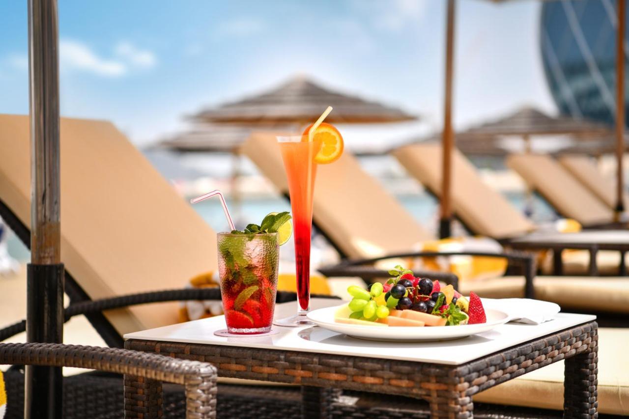Al Raha Beach Resort & Spa Abu Dabi Exterior foto The photo shows a table set outdoors, featuring two colorful cocktails and a plate of food. One cocktail is a vibrant red drink garnished with mint, while the other is a layered orange beverage with an orange slice on the rim. The plate of food inclu