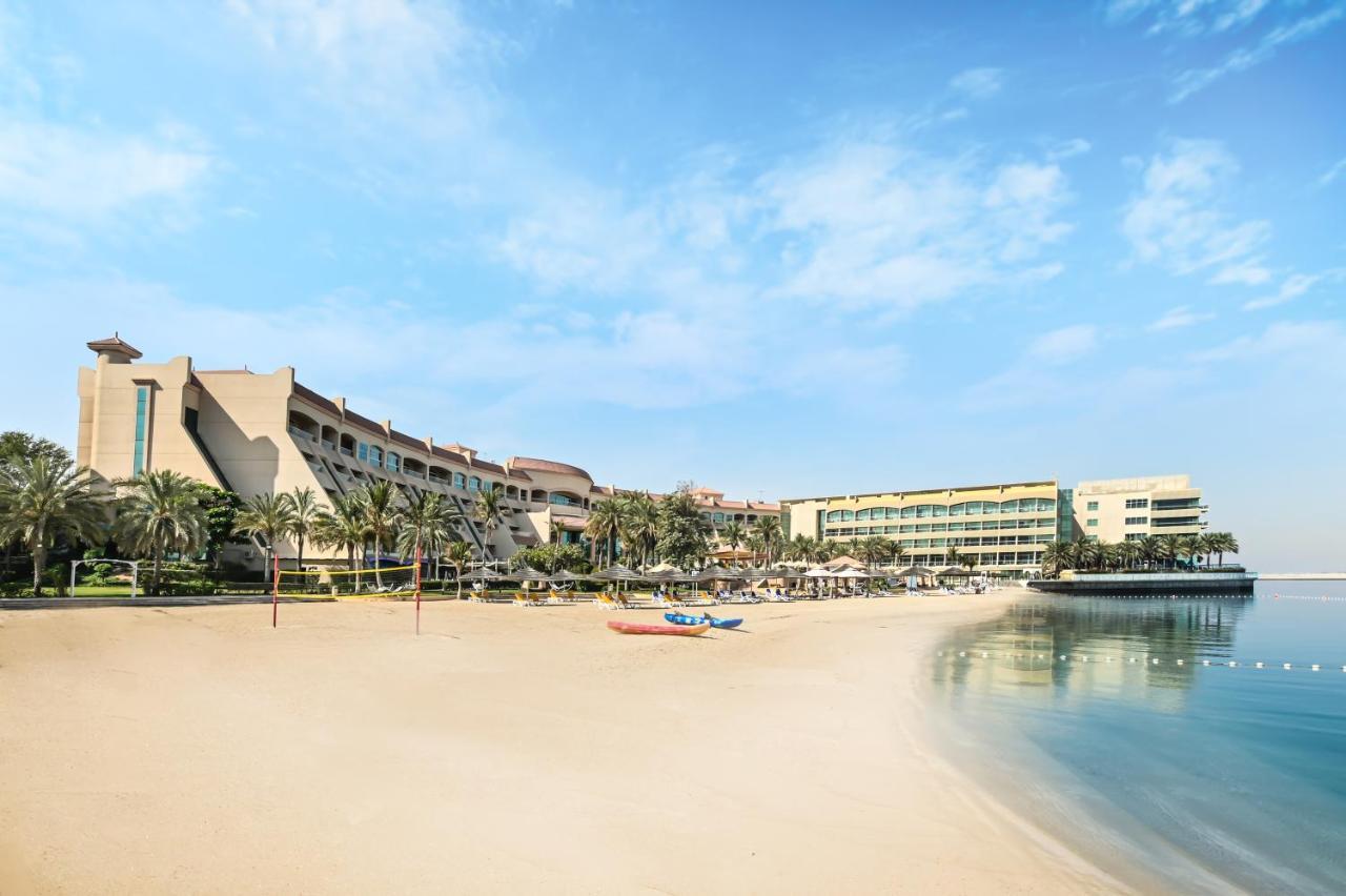 Al Raha Beach Resort & Spa Abu Dabi Exterior foto The photo depicts a serene beach scene with a clear blue sky. In the foreground, there is a sandy beach that gently slopes down to calm water. On the left side, several palm trees provide a tropical atmosphere. In the background, there are two large 