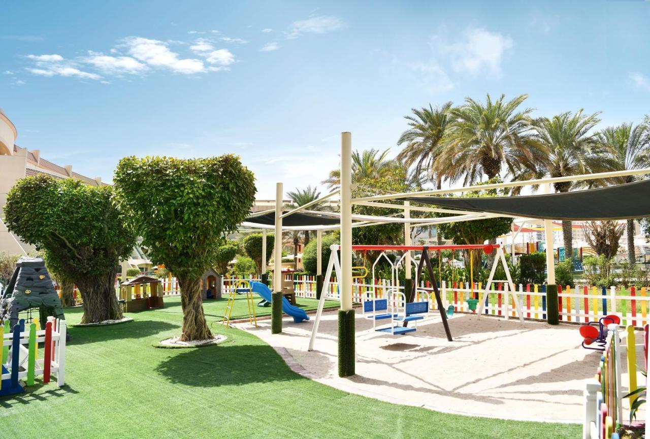 Al Raha Beach Resort & Spa Abu Dabi Exterior foto The photo shows a brightly lit outdoor playground surrounded by palm trees. The playground features a variety of play equipment, including swings, a slide, and a climbing structure. The ground is covered in lush green artificial grass, providing a so