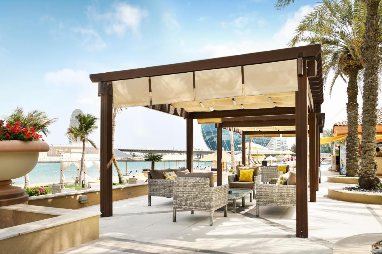 Al Raha Beach Resort & Spa Abu Dabi Exterior foto The photo shows a stylish outdoor seating area by a beach. There is a wooden pergola covered with a light fabric shade, providing shelter to a set of comfortable wicker chairs and tables. The area is adorned with palm trees, and in the background, th