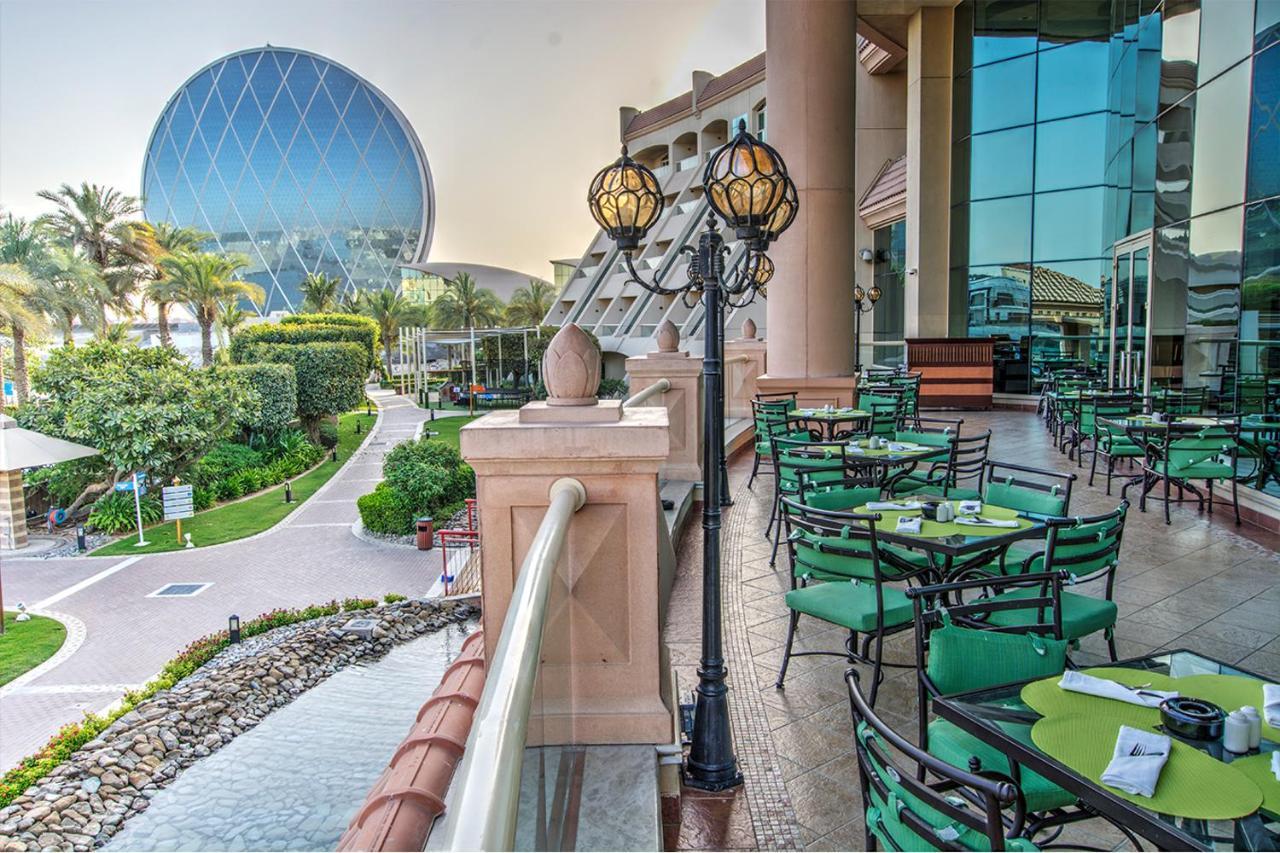 Al Raha Beach Resort & Spa Abu Dabi Exterior foto The photo depicts an outdoor dining area with green tables and chairs arranged on a patio. In the background, there is a large, distinctive building with a striking blue dome, surrounded by well-maintained landscaping, including trees and plants. The