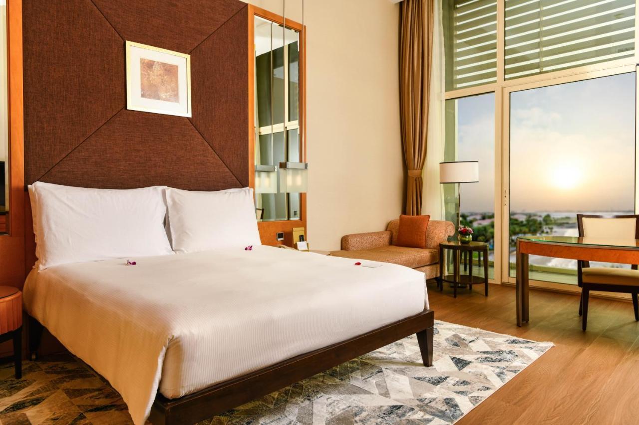 Al Raha Beach Resort & Spa Abu Dabi Exterior foto The photo shows a stylish hotel room featuring a large bed with white bedding, positioned against a textured wall. There’s a small seating area with a chair and a table, and a window with curtains that allows natural light to fill the space. The room