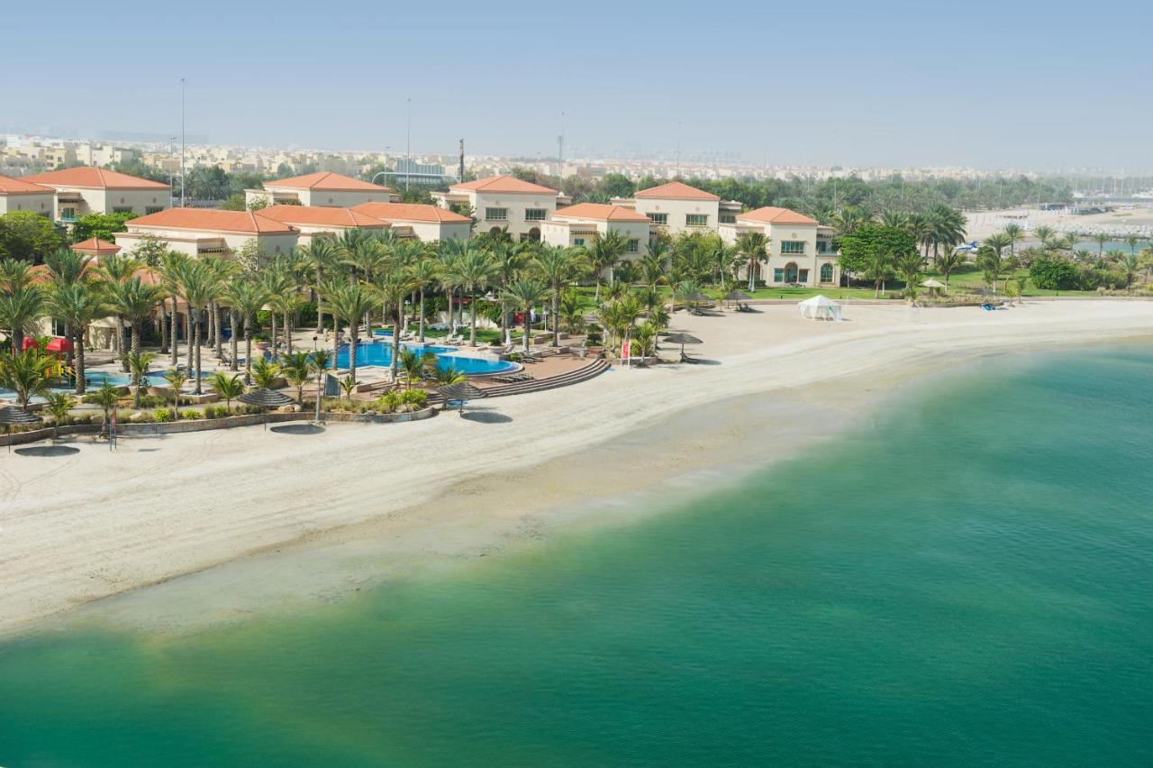 Al Raha Beach Resort & Spa Abu Dabi Exterior foto The photo depicts a serene beach scene with a clear turquoise sea and a sandy shoreline. In the background, there are several buildings featuring a Mediterranean architectural style with red-tiled roofs. The area is adorned with palm trees and outdoo