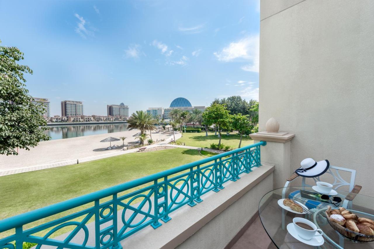 Al Raha Beach Resort & Spa Abu Dabi Exterior foto The photo shows a balcony view overlooking a scenic landscape. In the foreground, there is a glass table set with snacks or pastries and two cups. Beyond the balcony railing, there is a well-maintained green area with palm trees and a pathway. The ba