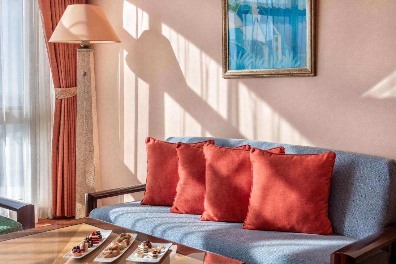 Al Raha Beach Resort & Spa Abu Dabi Exterior foto The photo shows a cozy interior setting, likely a sitting area. It features a light blue couch with four red cushions. In front of the couch, there is a glass table displaying an assortment of snacks or small dishes. The room has soft lighting, with 