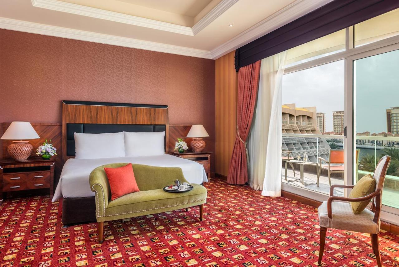 Al Raha Beach Resort & Spa Abu Dabi Exterior foto The photo shows a hotel room with an elegant and modern design. It features a large bed with white linens and a dark wooden headboard. On either side of the bed are matching bedside lamps. There is a green armchair with a red pillow positioned near t