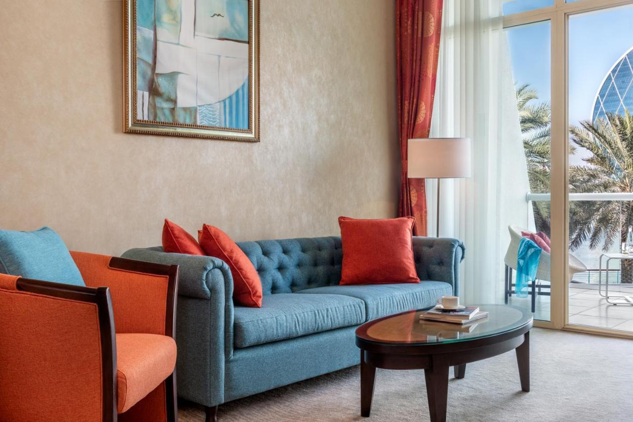 Al Raha Beach Resort & Spa Abu Dabi Exterior foto The photo shows a well-decorated living room. There is a plush blue couch with orange accent pillows and a cozy orange armchair nearby. A round coffee table sits in front of the couch, and above it, there is a colorful painting hanging on the wall. L