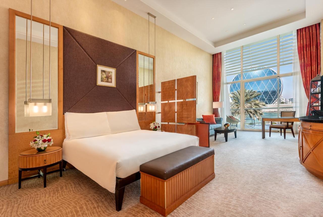 Al Raha Beach Resort & Spa Abu Dabi Exterior foto The photo shows a modern hotel room featuring a neatly made bed with a white comforter, situated against a stylish headboard with a geometric design. There are bedside tables on either side of the bed, each with a small flower arrangement. A comforta