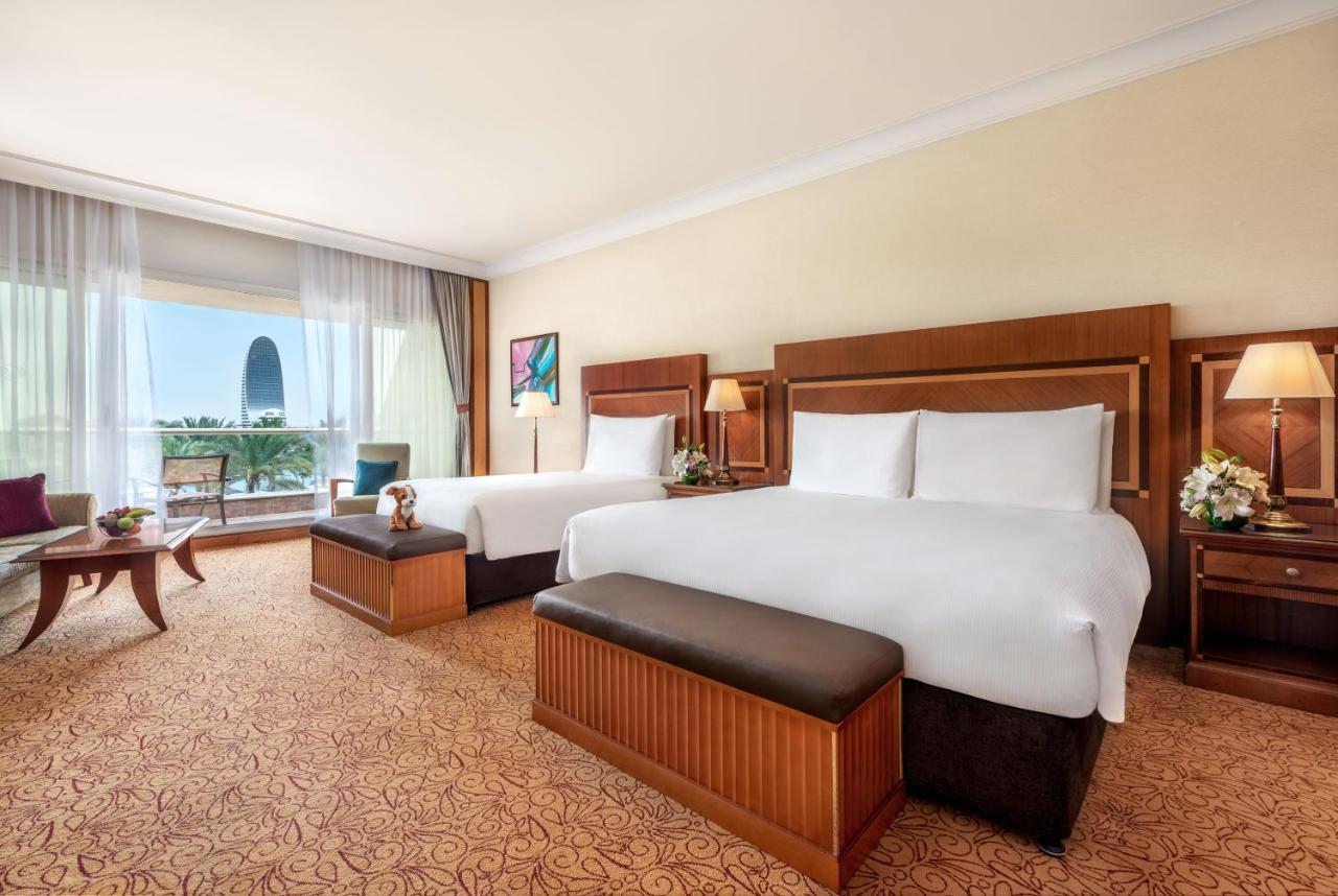 Al Raha Beach Resort & Spa Abu Dabi Exterior foto The photo shows a spacious hotel room featuring two large beds with white linens and dark brown upholstered headboards. There are matching benches at the foot of each bed. The room is decorated with a warm color palette and has a carpeted floor. To o
