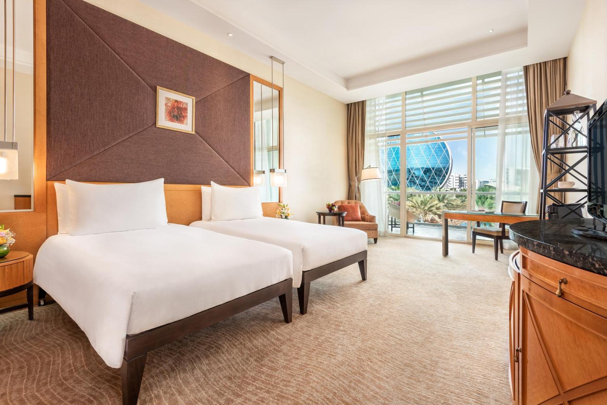 Al Raha Beach Resort & Spa Abu Dabi Exterior foto The photo shows a well-decorated hotel room featuring two single beds with white linens. The room has a modern aesthetic, with a large window letting in natural light and offering a view of a circular structure outside, possibly a landmark or artisti