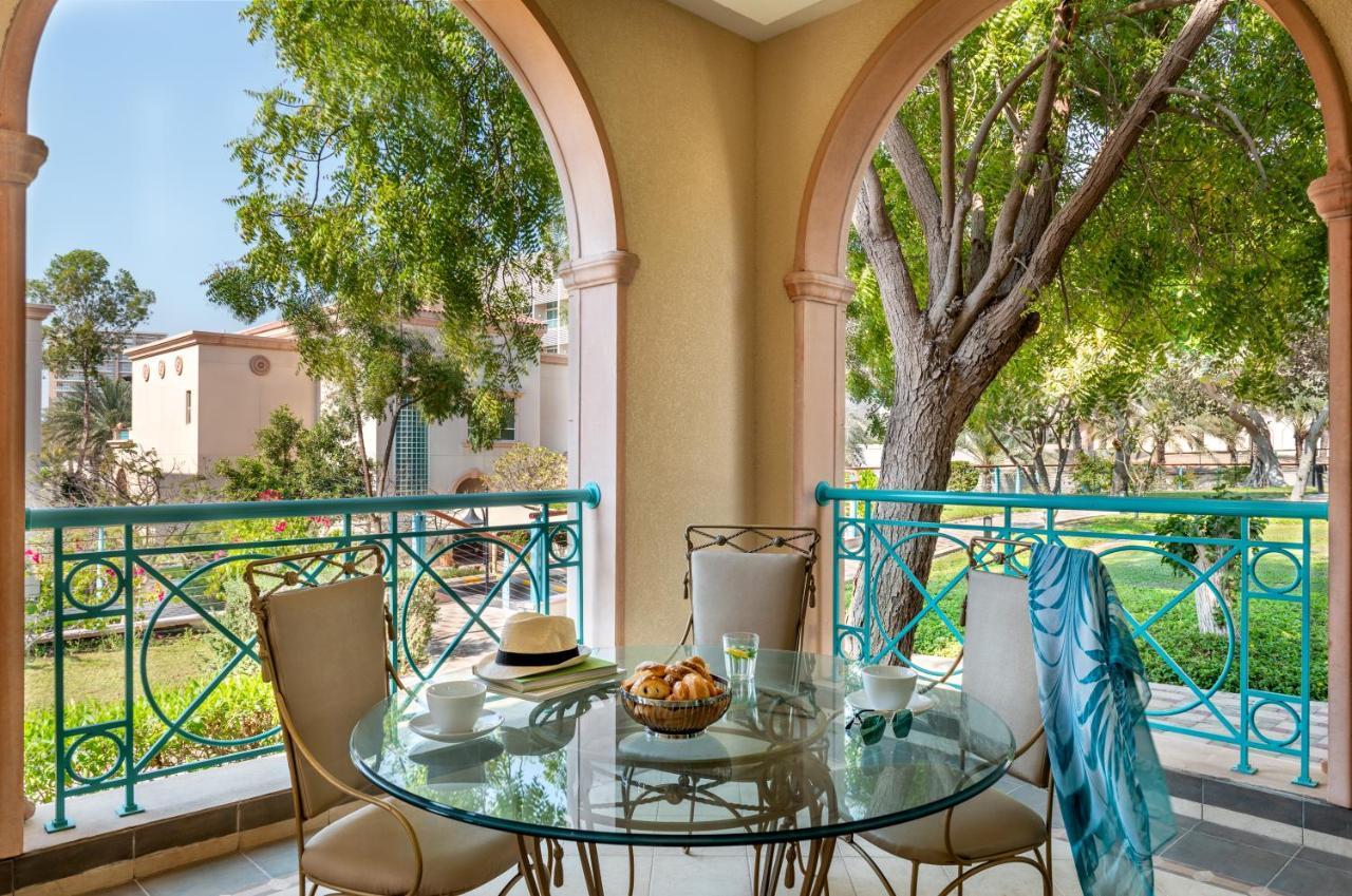 Al Raha Beach Resort & Spa Abu Dabi Exterior foto The photo shows a serene outdoor balcony setting. There is a circular glass table with a bowl of fruit or snacks in the center, alongside a glass and a small plate. Surrounding the table are four elegant chairs. The railing is turquoise, framing a vi