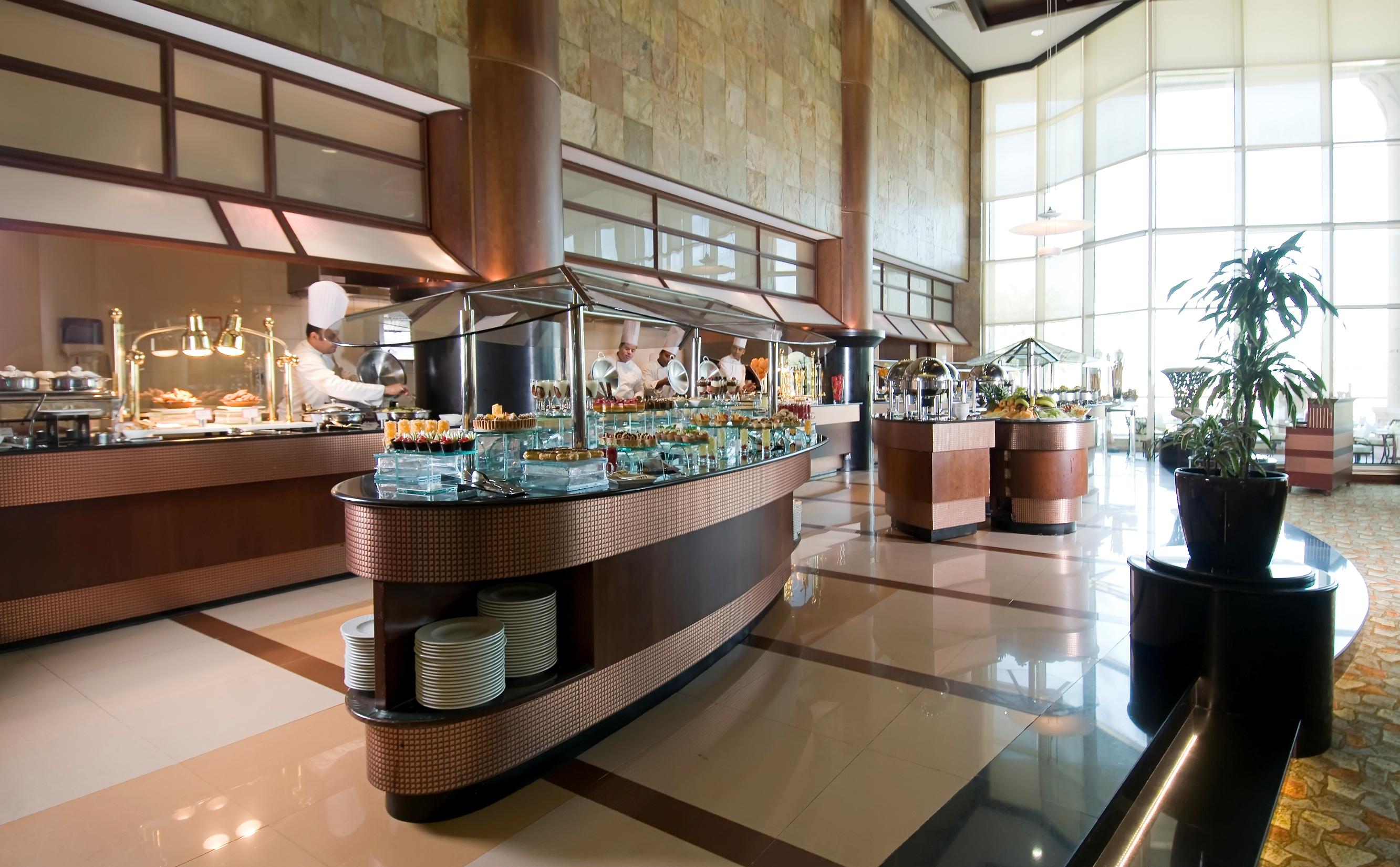 Al Raha Beach Resort & Spa Abu Dabi Exterior foto The photo shows a spacious buffet area in a restaurant or hotel. There are multiple food stations with glass displays showcasing a variety of dishes. Chefs can be seen in the background, preparing food. The decor features large windows allowing natur