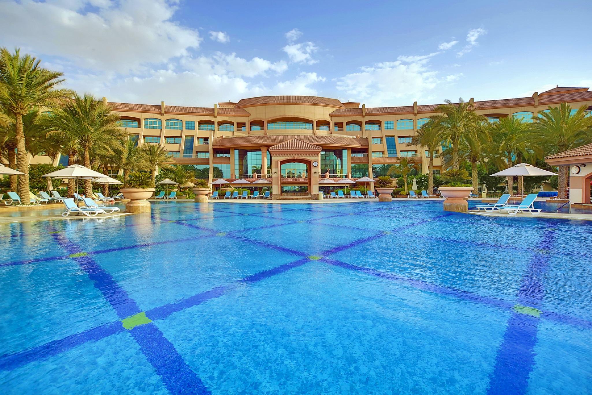 Al Raha Beach Resort & Spa Abu Dabi Facilidades foto The photo features a luxurious resort with a large swimming pool in the foreground. The pool is crystal clear and reflects the sky, bordered by lounge chairs and palm trees. In the background, the resort building showcases modern architecture with la