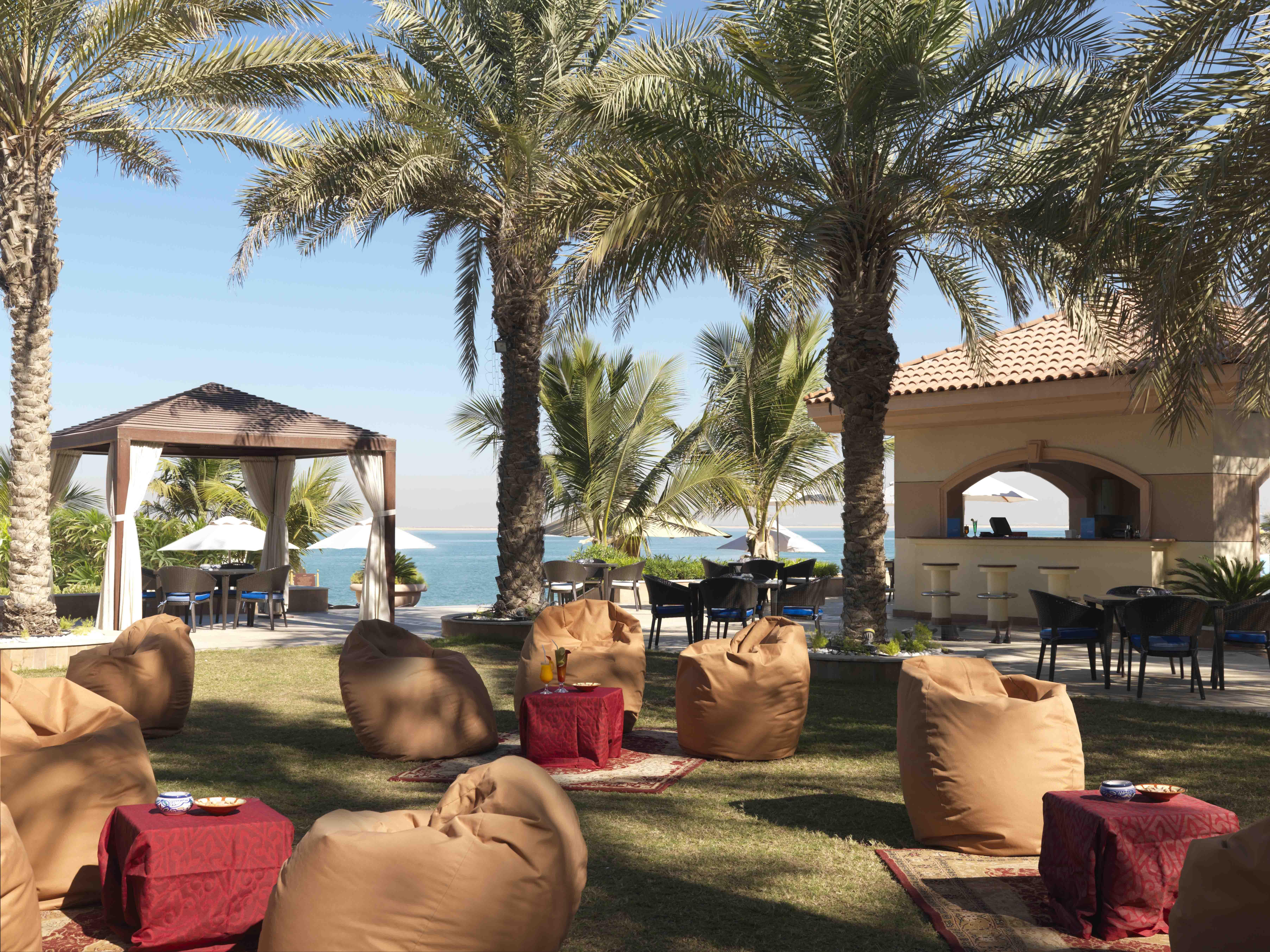 Al Raha Beach Resort & Spa Abu Dabi Restaurante foto The photo depicts a beautiful outdoor setting with several elements of relaxation and leisure. In the foreground, there are large, comfortable bean bag chairs arranged on a grassy area, accompanied by low tables. Surrounding this seating area are tal