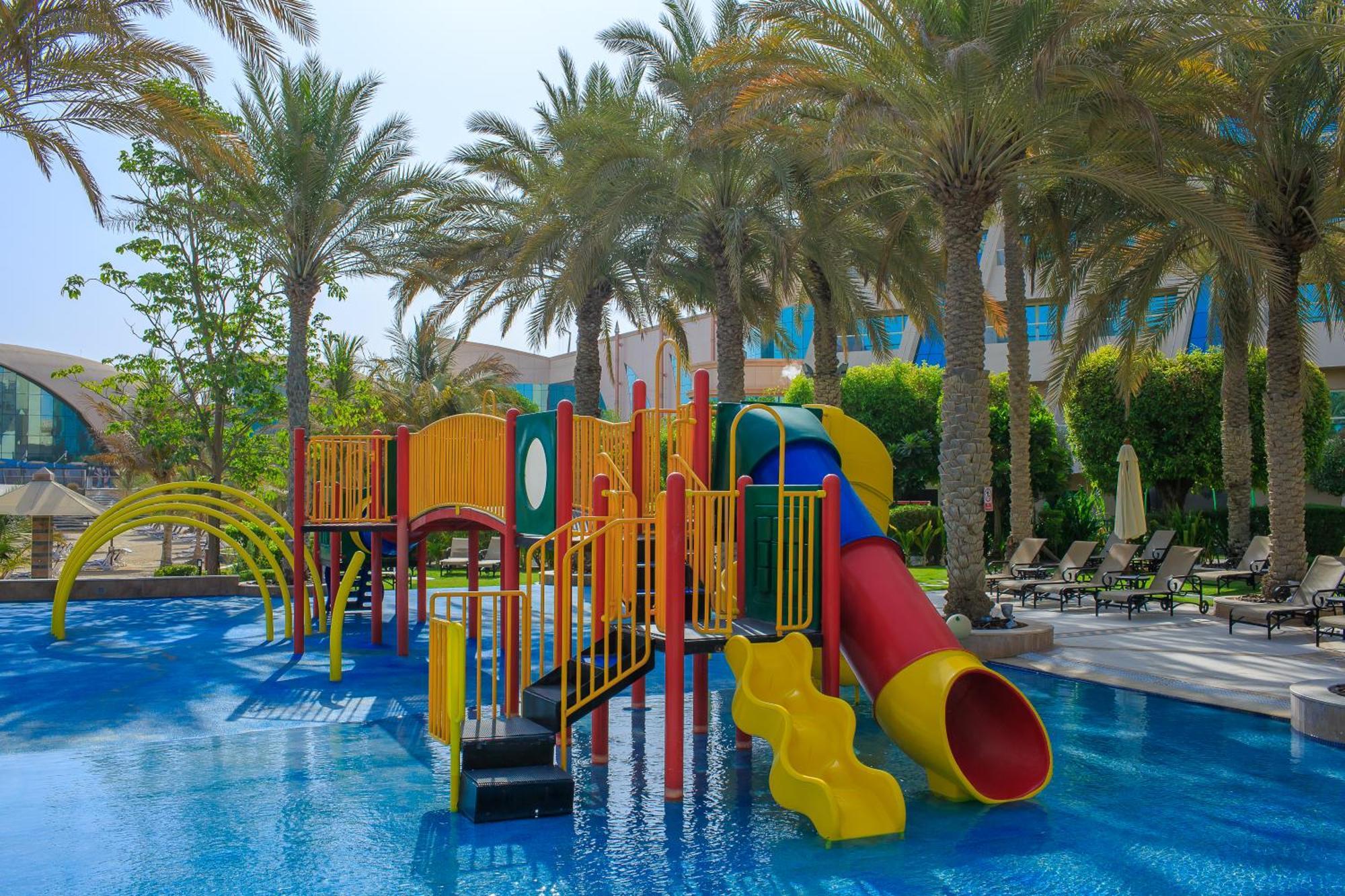Al Raha Beach Resort & Spa Abu Dabi Exterior foto The photo shows a colorful water play area located in a pool setting. It features a variety of play structures, including slides in multiple colors, such as yellow, red, and blue. Surrounding the play structure are palm trees, creating a tropical atm