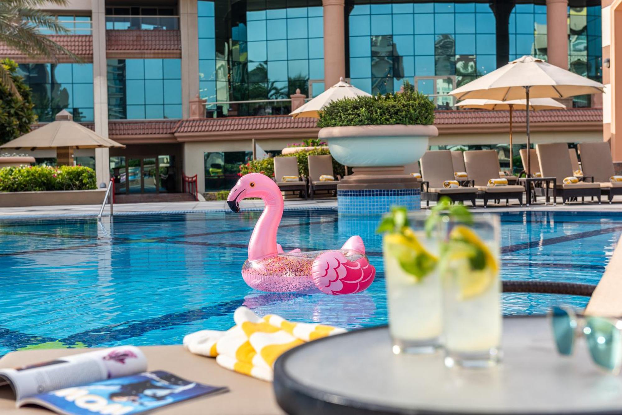 Al Raha Beach Resort & Spa Abu Dabi Exterior foto The photo depicts a bright and inviting outdoor swimming pool surrounded by a luxurious setting. In the pool, there is a large pink flamingo float, adding a fun and whimsical touch. To the side, a table is set up with two refreshing drinks, likely co