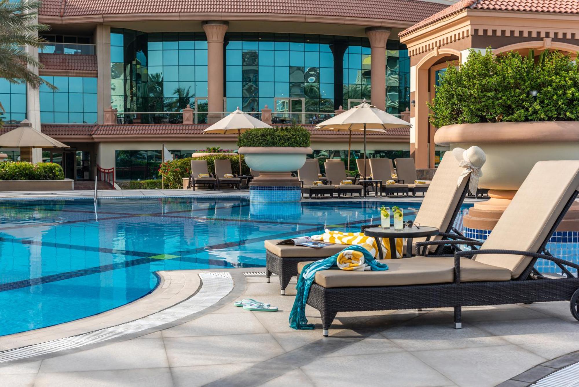 Al Raha Beach Resort & Spa Abu Dabi Exterior foto The photo depicts a luxurious outdoor swimming pool area. In the foreground, there are two lounge chairs with cushions, positioned beside the pool. A small table between them holds drinks and snacks, suggesting a relaxing atmosphere. The pool itself 