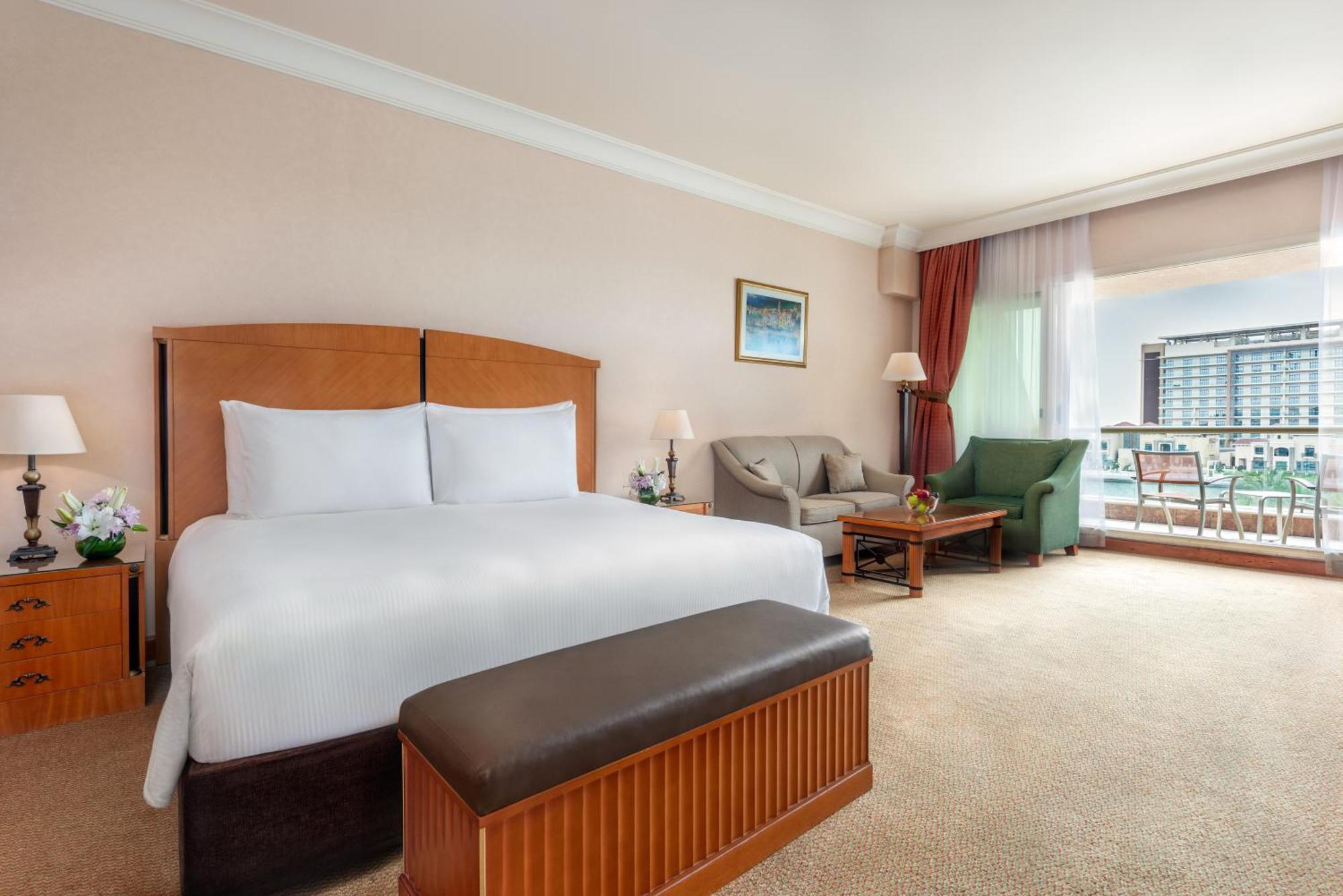 Al Raha Beach Resort & Spa Abu Dabi Exterior foto The photo shows a well-furnished hotel room featuring a large bed with white linens and a wooden headboard. There's a brown bench at the foot of the bed. To one side, a sitting area includes a sofa and a coffee table, complemented by two lamps. Large