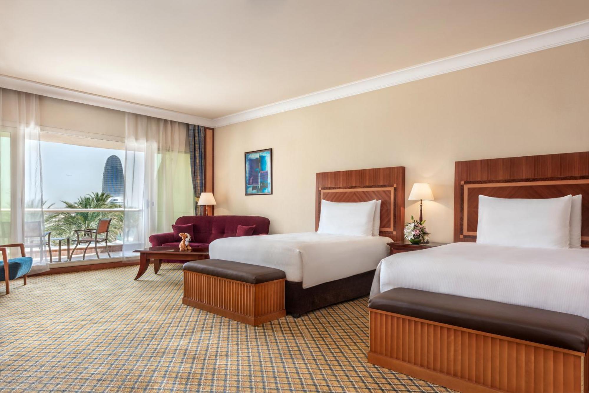 Al Raha Beach Resort & Spa Abu Dabi Habitación foto The photo shows a hotel room featuring two twin beds with wooden headboards. The room has a light, neutral color scheme, with a large window that provides natural light and a view of the outdoors. There is a small seating area with a purple sofa and 