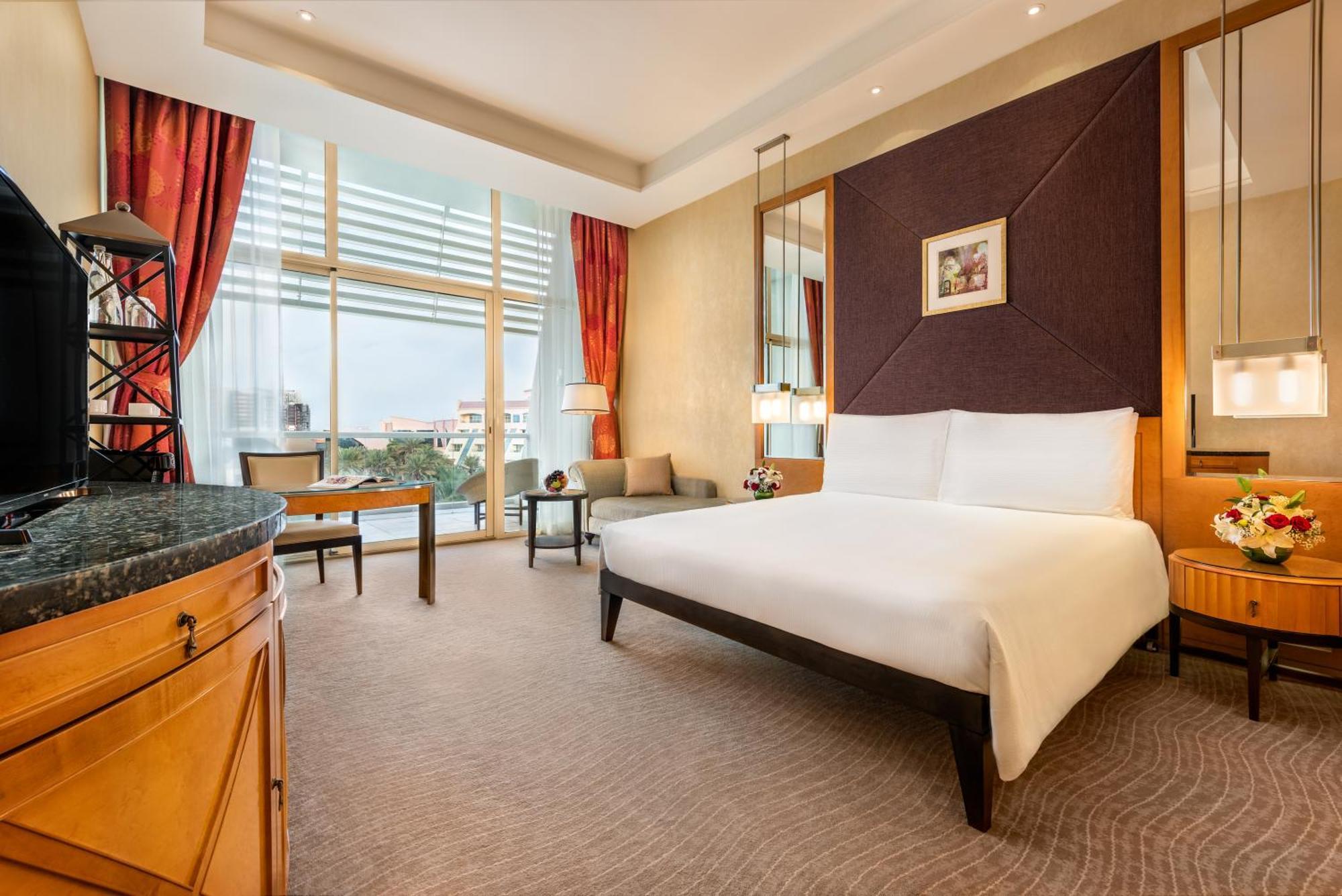 Al Raha Beach Resort & Spa Abu Dabi Habitación foto The photo shows a modern hotel room featuring a large bed with a white duvet positioned against a purple accent wall. There are tall windows with sheer curtains allowing natural light to enter the space, along with red drapes framing the windows. A d
