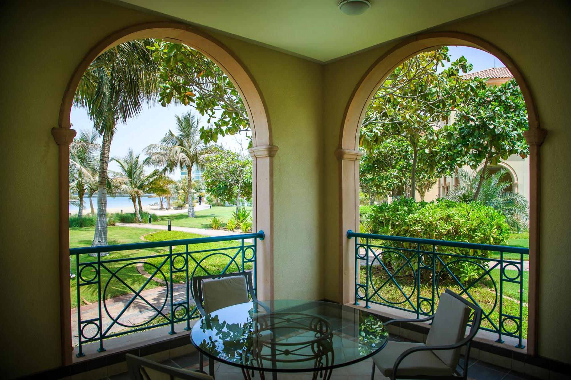 Al Raha Beach Resort & Spa Abu Dabi Habitación foto The photo displays a beautifully designed balcony or terrace area. It features two large arches that frame the view outside. Through the arches, you can see a lush garden with palm trees and well-maintained greenery. There’s a glass table with severa