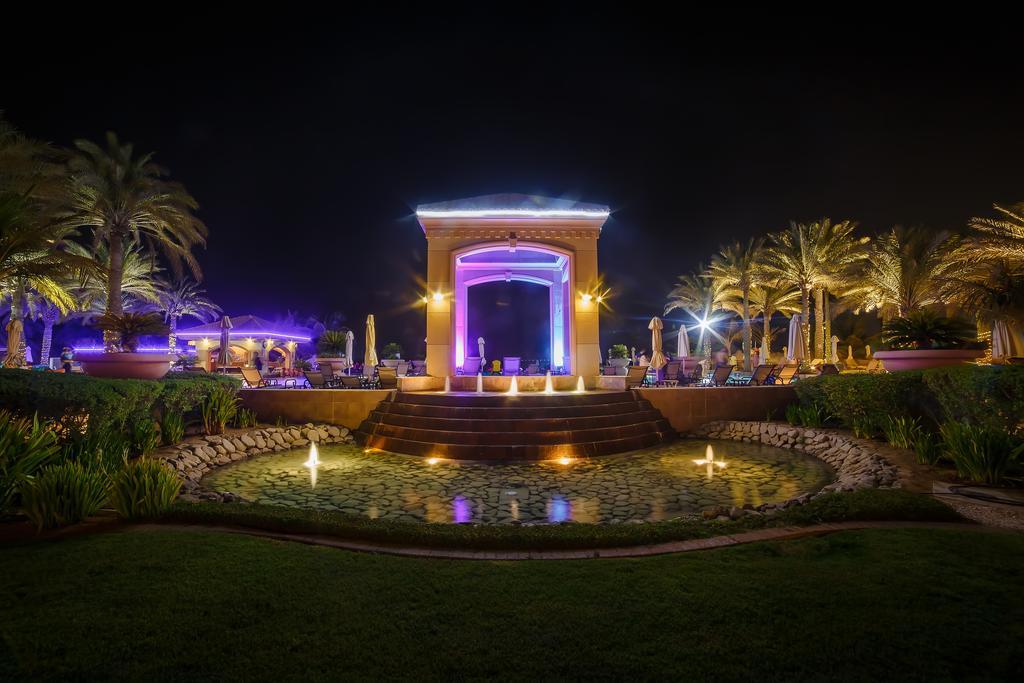 Al Raha Beach Resort & Spa Abu Dabi Exterior foto The photo depicts a beautifully lit outdoor scene at night. In the foreground, there is a serene water feature with a circular pond, and small fountains creating gentle ripples. Surrounding the pond are lush green lawns and ornamental plants. 

In th