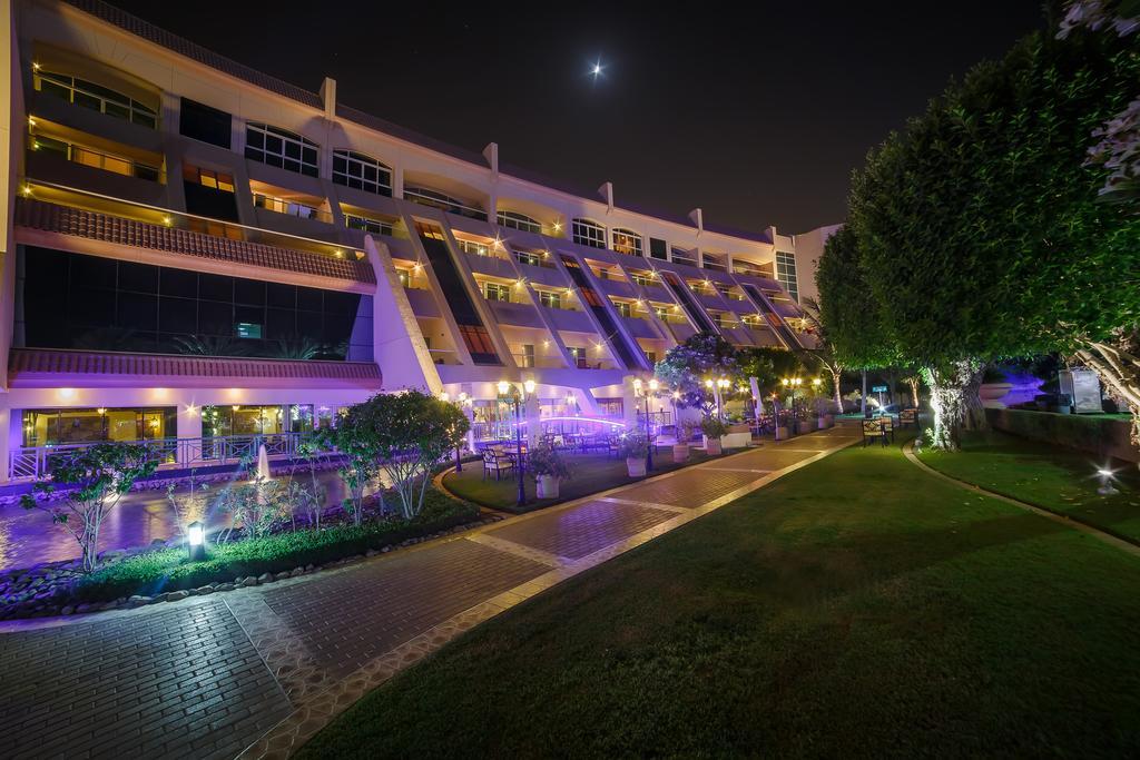 Al Raha Beach Resort & Spa Abu Dabi Exterior foto The photo shows a modern building illuminated at night, with a vibrant purple hue. The structure has multiple floors, each with balconies overlooking a landscaped area. In the foreground, there is a pathway lined with well-manicured grass and shrubs,