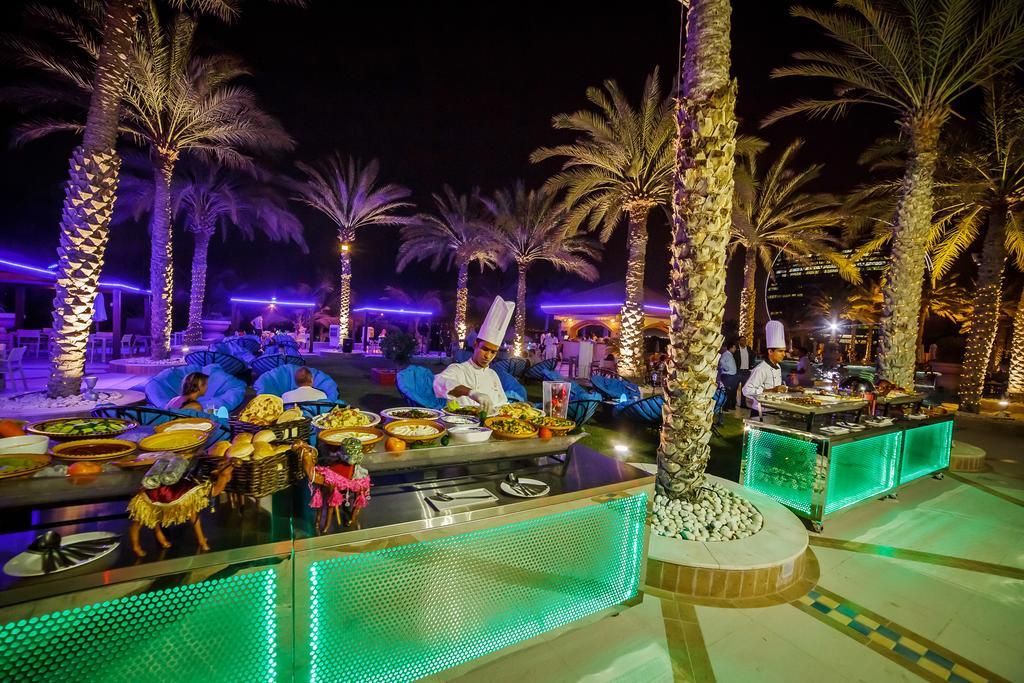 Al Raha Beach Resort & Spa Abu Dabi Exterior foto The photo depicts an outdoor dining area at night, featuring several chefs in white uniforms and hats working at a food station. The setting is illuminated by colorful lights, with glowing green accents on the serving tables. Surrounding the area are